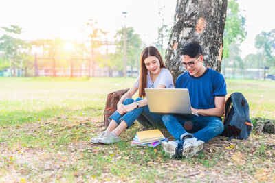 Friends studying over laptop while sitting by tree at park