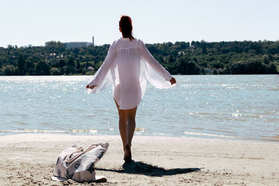 Rear view of carefree woman in white shirt walking on a sand at the beach.