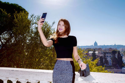 Smiling woman taking selfie through mobile phone against sky