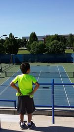 Rear view of boy holding tennis balls while standing by railing at court