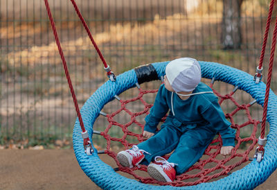 Cute little boy with down syndrome in a funny hat walks in the playground, swinging on a swing