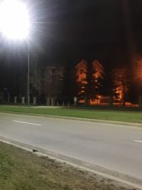 Empty road by illuminated buildings against sky at night