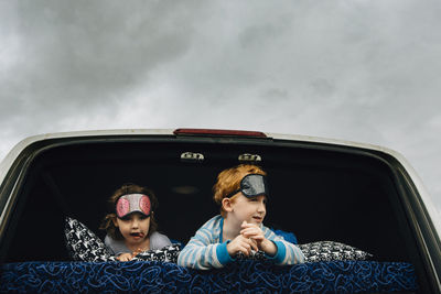 Siblings with sleep masks lying in vehicle while looking away against cloudy sky