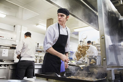 Male chef student tossing meat with colleagues in background at cooking school