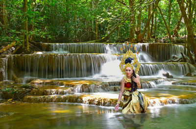 Portrait of beautiful young woman wearing crown and traditional clothing while sitting against waterfall in forest