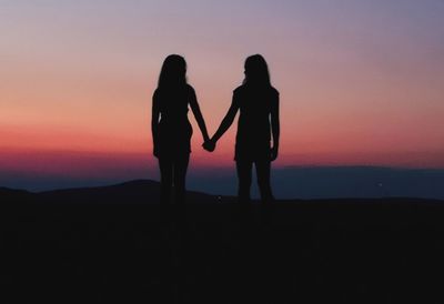 Silhouette girls holding hands while standing against sky during sunset