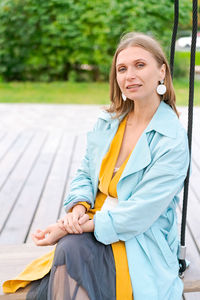 Happy woman swinging on swing in city park, wearing yellow dress and blue coat