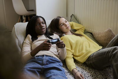 Teenage girl playing video game while lying down with female friend in bed at home