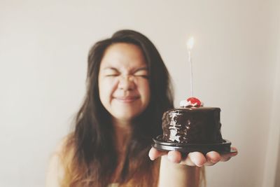 Woman making face while holding birthday cake against wall