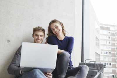 Smiling young couple sitting at concrete wall sharing laptop