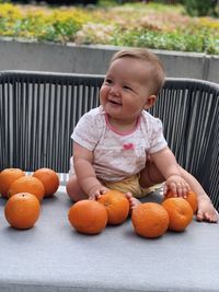 Blond baby girl sitting wearing a light pink shirt and sitting on a table with big oranges around 