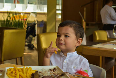 Cute boy eating french fries while sitting on chair at restaurant