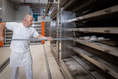 Side view of male worker in workwear putting raw dough on flat shovel into electric oven while working in bakery