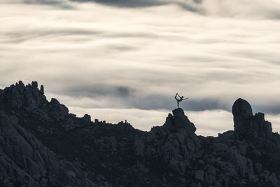 Silhouette of unrecognizable person doing yoga in graceful pose standing on peak of rough cliff with colorful clouds on background