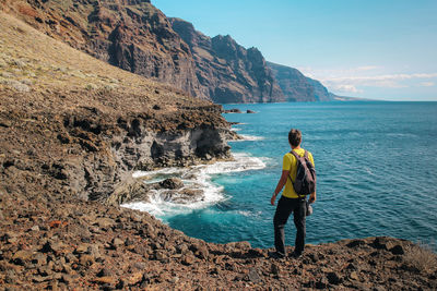 Rear view of man with a backpack standing on rock by sea against sky