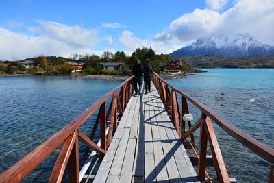 Rear view of people walking on footbridge over lake against sky