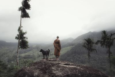 Rear view of monk with dog standing on mountain peak against clear sky