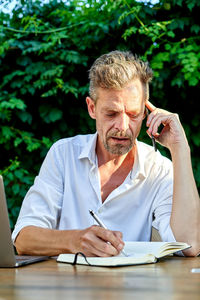Glad mature businessman in white shirt smiling and reading notes in planner while sitting at table near bushes and answering phone call during work in garden