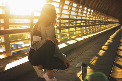 Full length of female athlete with bottle crouching on footbridge during sunset