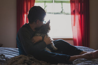 Boy embracing dog while sitting on bed at home