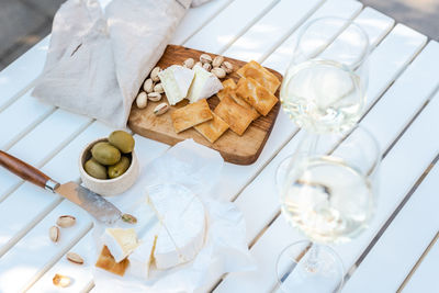 Two glasses of white wine and a wooden plate with cheese and nuts on a white table outdoors.