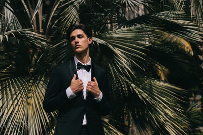 A beautiful young man, the groom in an elegant wedding suit, stands posing in the city's old park