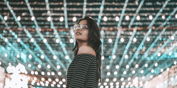 Portrait of beautiful young woman standing against illuminated wall