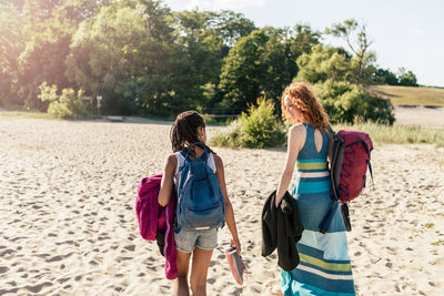 Rear view of mother and daughter with backpacks walking at beach on sunny day