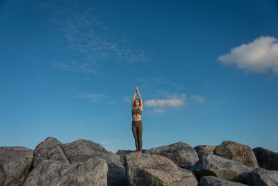 Young female in warm activewear standing in mountain pose with arms up while practicing yoga on rock against blue sky