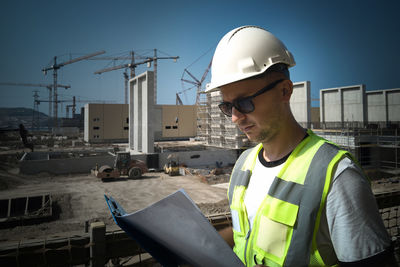 Caucasian civil engineer dressed in white hardhat and protective vest looks at paperwork