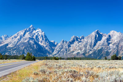 Road by field leading towards snowcapped mountains against clear blue sky