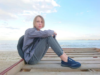 Portrait of young woman sitting on shore at beach against sky