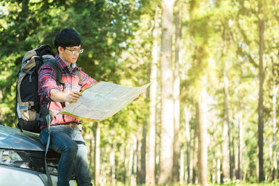 Male hiker holding map while sitting on car hood against trees