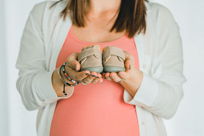 Midsection of pregnant woman holding baby shoes against white background