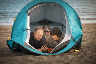 Couple using mobile phone while resting with dog in tent at beach