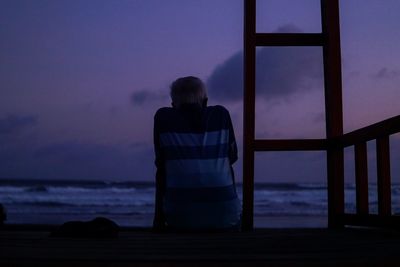 Rear view of man sitting by beach against sky at dusk