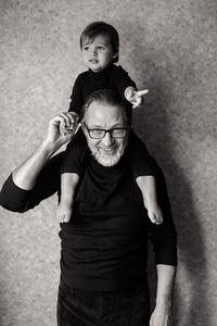 Grandfather with beard and glasses with his grandson sitting on the neck in the studio near the wall