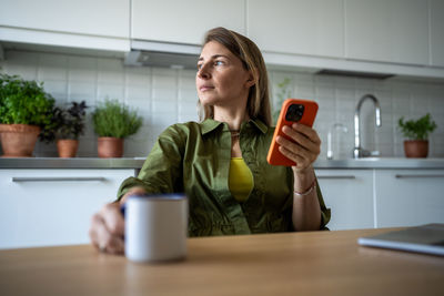 Thoughtful woman sits in kitchen with cup holding smartphone immersed daydreaming looking to window.