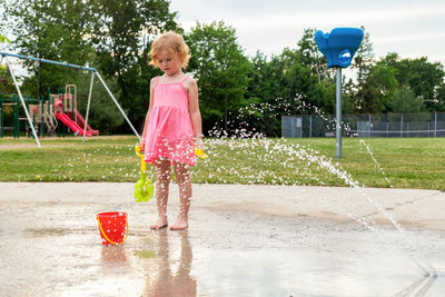  small beautiful girl in pink dress having fun at fountain playground in park in summer