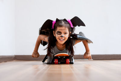 Portrait of cute playful girl with spooky make-up lying on skateboard at home during halloween