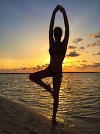 Young woman with tree pose at beach against sky during sunset