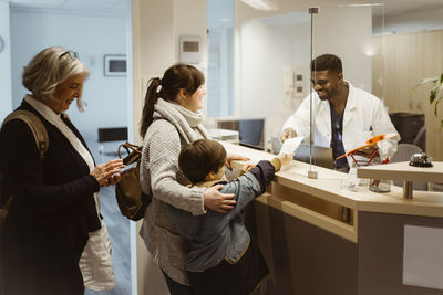 Boy passing form to male receptionist below transparent shield in clinic