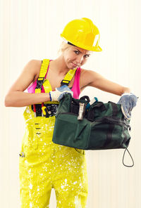 Portrait of female worker in coverall holding tool bag while standing against wall