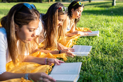 Side view of young women lying on grass while reading book at park