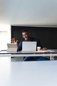 Positive african american adult male in formal clothes and eyeglasses sitting at table while having video call on tablet near netbook and documents with takeaway drink in bright office