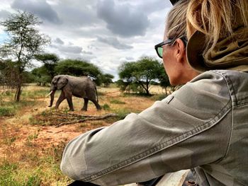 Woman looking at elephant walking on grass in forest against sky