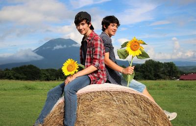 Boys holding sunflowers while sitting on hay bale against sky