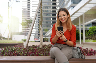 Businesswoman sitting relaxed on bench outside office using her smartphone. 
