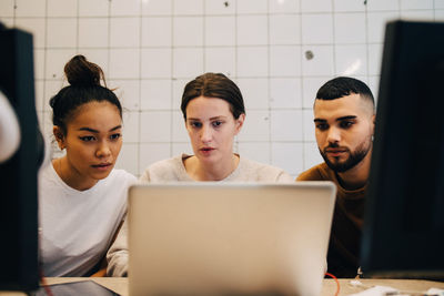 Young multi-ethnic computer hackers looking at laptop against wall in office