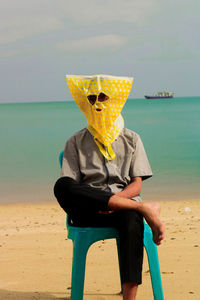 Young man wearing yellow plastic bag while sitting on chair at beach against sky